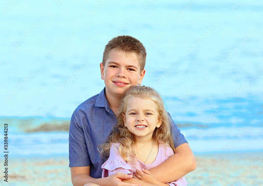 Beautiful Caucasian kids are sitting on the beach. A boy in a blue shirt, girl in a pink dress. Rest and travel. Family day. Happy family concept. 
People and advertising concept