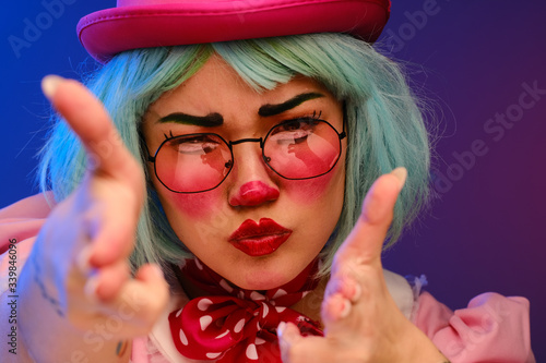 Close-up portrait of a clown girl with blue hair in a pink hat, pink dress and glasses. A clown shows different human emotions. 