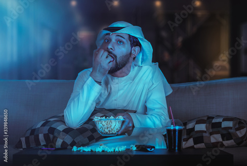 front View of an Arab Man Sitting on a Couch Watching Movie. Arabic Man watching fascinating program in TV and eating popcorn.