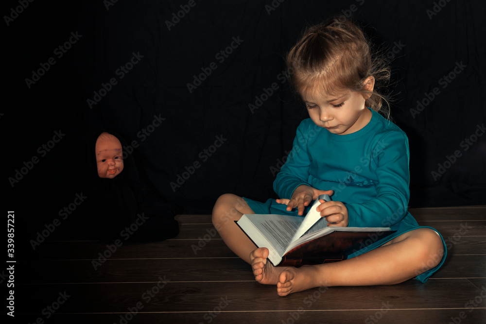 a little girl in a blue dress 4 years old is sitting on the floor in a dark room and reading a book to a doll. home schooling in quarantine and self-isolation due to coronavirus. dark key a little girl in a blue dress 4 years old is sitting on the floor in a dark room and reading a book to a doll. home schooling in quarantine and self-isolation due to coronavirus. dark key