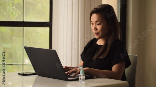 Asian lady working from home avoiding social distancing to fight coronavirus. Focus working on laptop with internet. Hand gel sanitizer on the table.