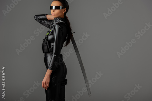side view of futuristic african american woman in glasses with sword on grey