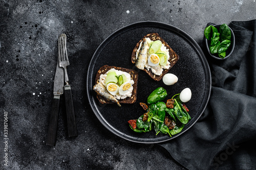 Sandwiches with sardines, egg, cucumber and cream cheese, salad garnish with spinach and dried tomatoes. Black background. Top view