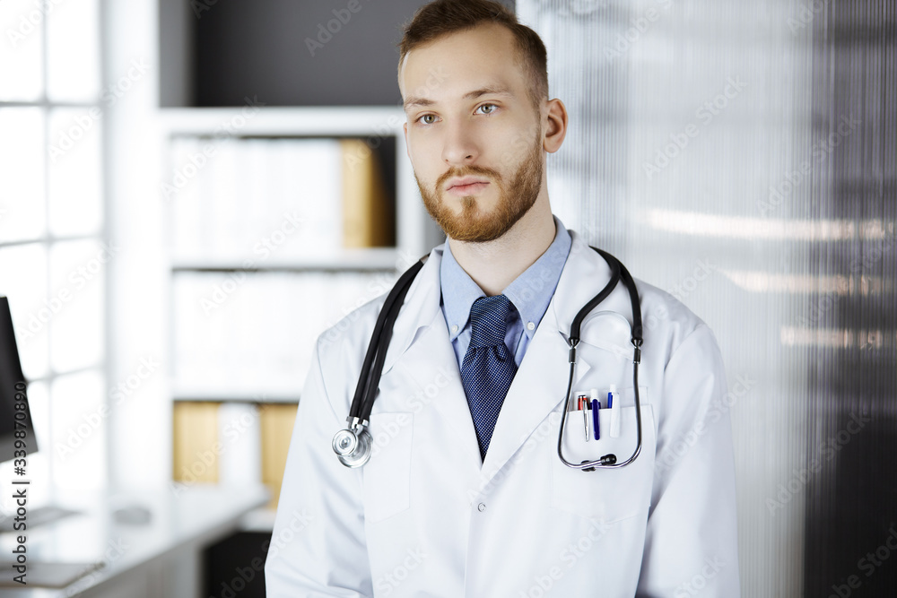 Red-bearded doctor standing straight in clinic near his working place. Portrait of physician. Medicine and healthcare concept Red-bearded doctor standing straight in clinic near his working place. Portrait of physician. Medicine and healthcare concept
