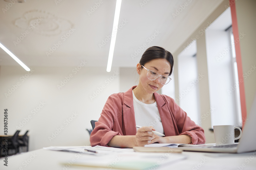 Young Asian businesswoman in eyeglasses sitting at her workplace and writing in documents at office Young Asian businesswoman in eyeglasses sitting at her workplace and writing in documents at office