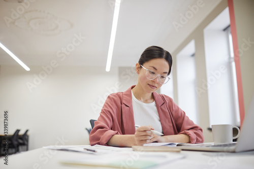Young Asian businesswoman in eyeglasses sitting at her workplace and writing in documents at office