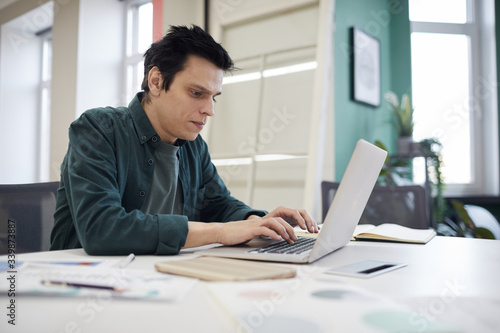 Young businessman in casual clothing sitting at his workplace and typing on laptop computer at office