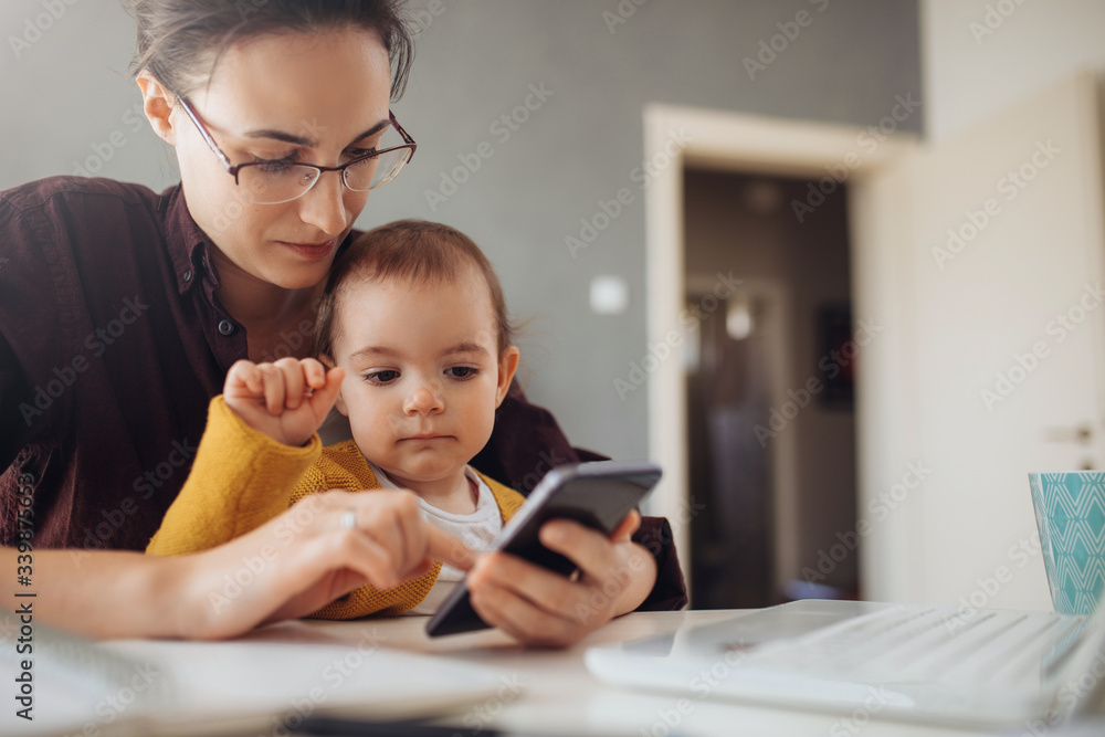 Young businesswoman working from home. Young mother using her mobile phone at her home office. She working with her child. Young businesswoman working from home. Young mother using her mobile phone at her home office. She working with her child.