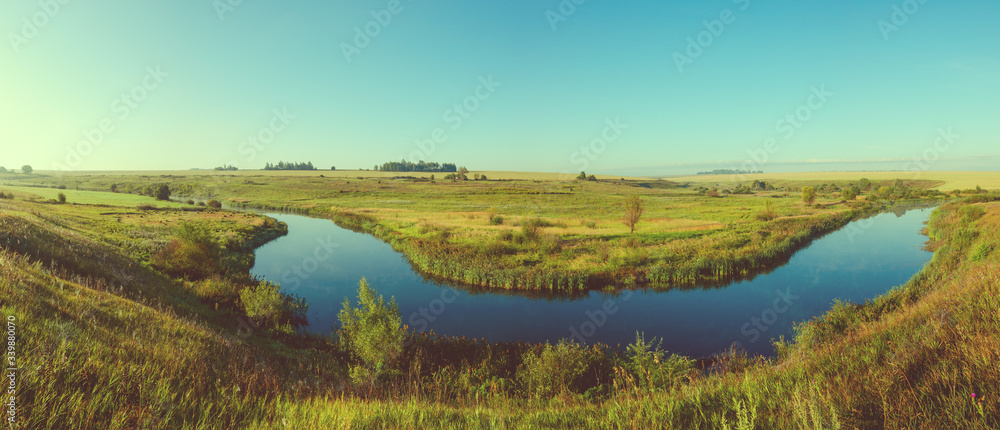 Sunny panoramic view of blue river and green meadows with trees on background.Retro and vintage colors. 
