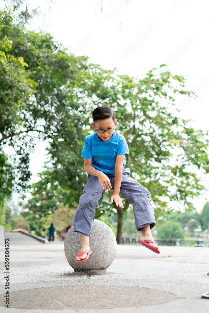 Outdoor portrait of a cute Malaysian little boy trying to jump from a concrete ball at the park.