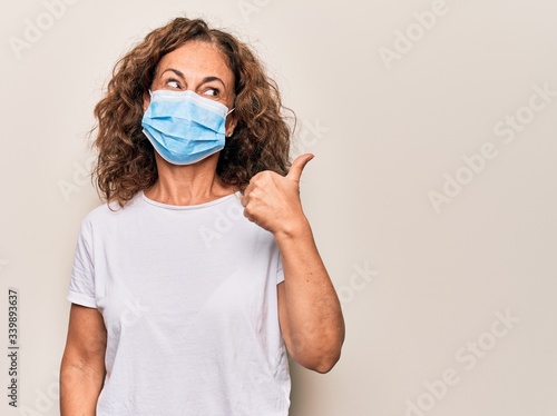 Middle age woman wearing coronavirus protection mask for covid-19 epidemic virus pointing thumb up to the side smiling happy with open mouth