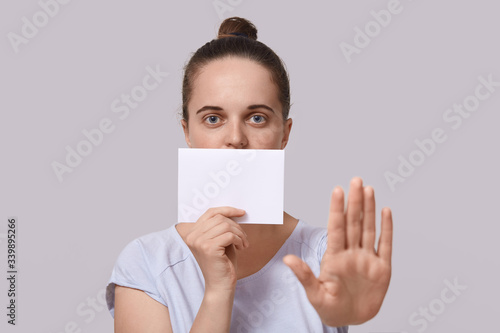 Picture of attractive female with hair bun, dresses casual white t shirt, looking directly at camera, posing against studio wall, covers mouth with sheet of paper and showing stop gesture with palm.