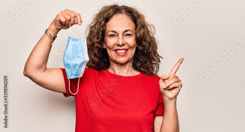 Middle age woman holding coronavirus protection mask for covid-19 epidemic virus smiling happy pointing with hand and finger to the side