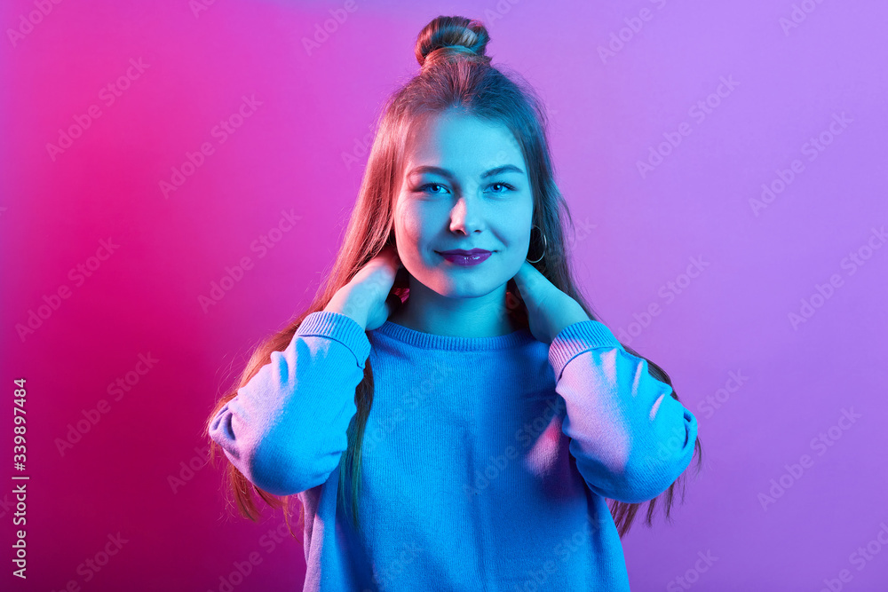 Fototapeta premium Indoor shot of attractive female wearing casual sweater, looking directly at camera, keeping both hands on neck, posing with hair bun isolated over pink neon background, lady has satisfied expression.