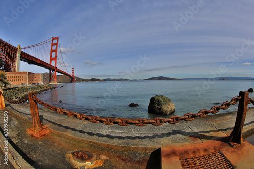 Golden Gate Bridge with weathered harsh rusty chain in foreground contrasting calm natural bay in background.