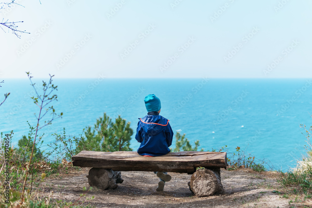 A lonely child is resting on a bench in the forest with a view of the sea.