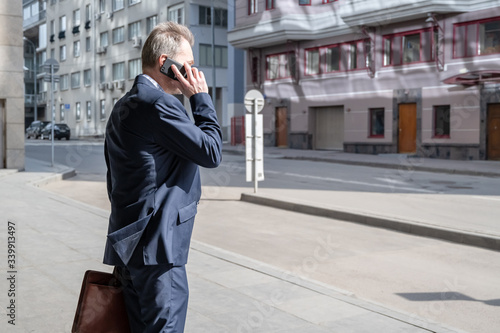 Alone middle-aged gray-haired businessman in a blue suit calling by phone standing on the empty city street.