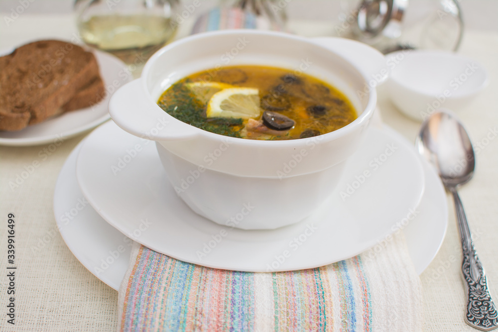 Traditional Russian meat soup solyanka with sausages, lemon and olives in a white ceramic bowl. Traditional Russian meat soup solyanka with sausages, lemon and olives in a white ceramic bowl.