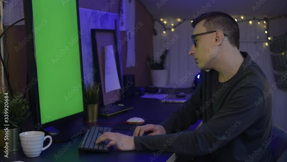 The guy at home sits at the computer and works or plays games behind a blue, green screen. The guy at home works as a freelancer and speaks on the phone, smiles, laughs, rejoice. The guy at home sits at the computer and works or plays games behind a blue, green screen. The guy at home works as a freelancer and speaks on the phone, smiles, laughs, rejoice.