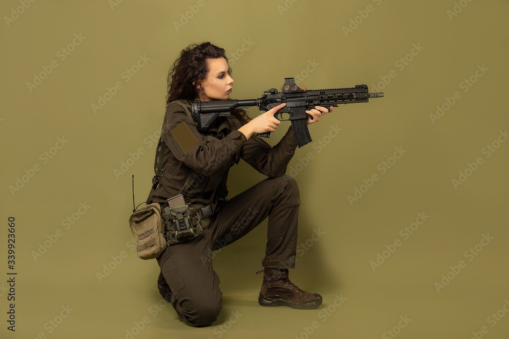 Military woman in green uniform with a vest unloading with a machine gun in her hands on a khaki background. A soldier sits with a gun Military woman in green uniform with a vest unloading with a machine gun in her hands on a khaki background. A soldier sits with a gun
