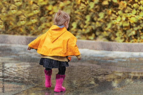 A happy little girl in a yellow raincoat and pink rubber boots walking in the rain on the street alone. Park, nature, outdoors. Childhood concept. Universal Children's Day.