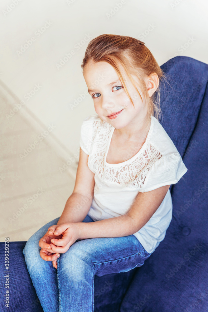 Stay Home Stay Safe. Sweet little girl in jeans and white T-shirt sitting on modern cozy blue chair relaxing in white bright living room at home indoors. Childhood schoolchildren youth relax concept.