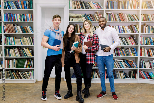 Group of four confident smiling multiethnical students, girls and boys in casual wear, posing in the library, holding books and looking at camera. Learning and education concept photo