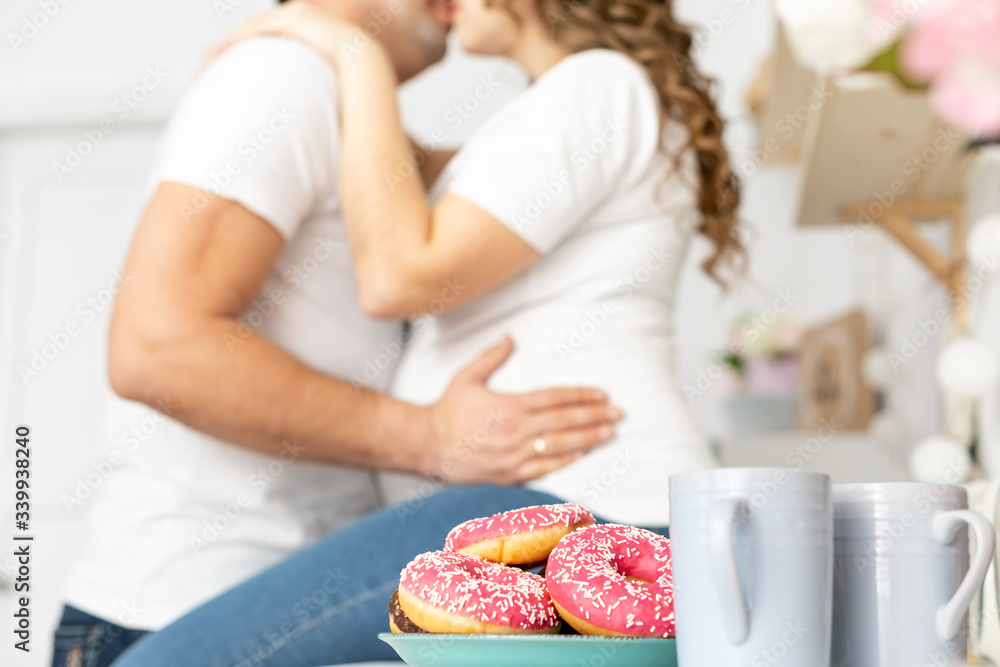 Man holding belly of his pregnant wife and kisses, sitting on the table in the kitchen with donuts in front