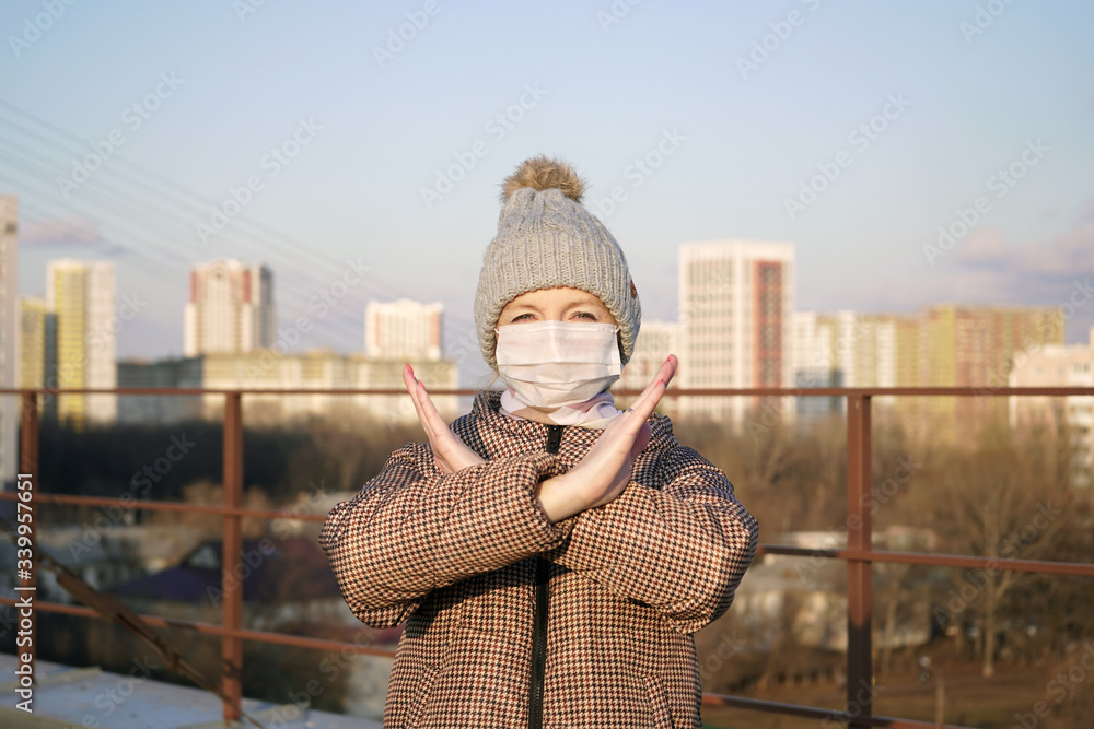 Young woman in a warm jacket and cap in a medical mask on the street crosses her arms in front of her against the background of the city. coronovirus, covid-19.