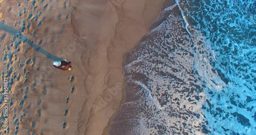 Aerial dinamic view from a top to a woman walking along the sea, next to the water, the seashore is full of human traces in the sand, looking at the rough sea, where huge foamy waves