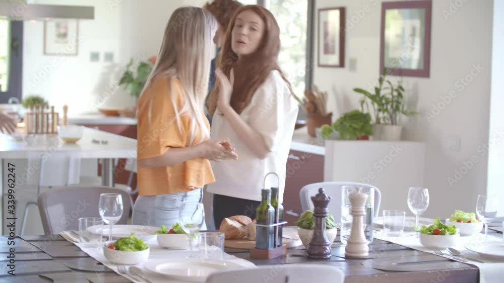 Young woman setting the table in living room putting water carafe for meal while happy friends talking.Happy people group enjoy modern home party dinner or lunch.Handheld video Young woman setting the table in living room putting water carafe for meal while happy friends talking.Happy people group enjoy modern home party dinner or lunch.Handheld video