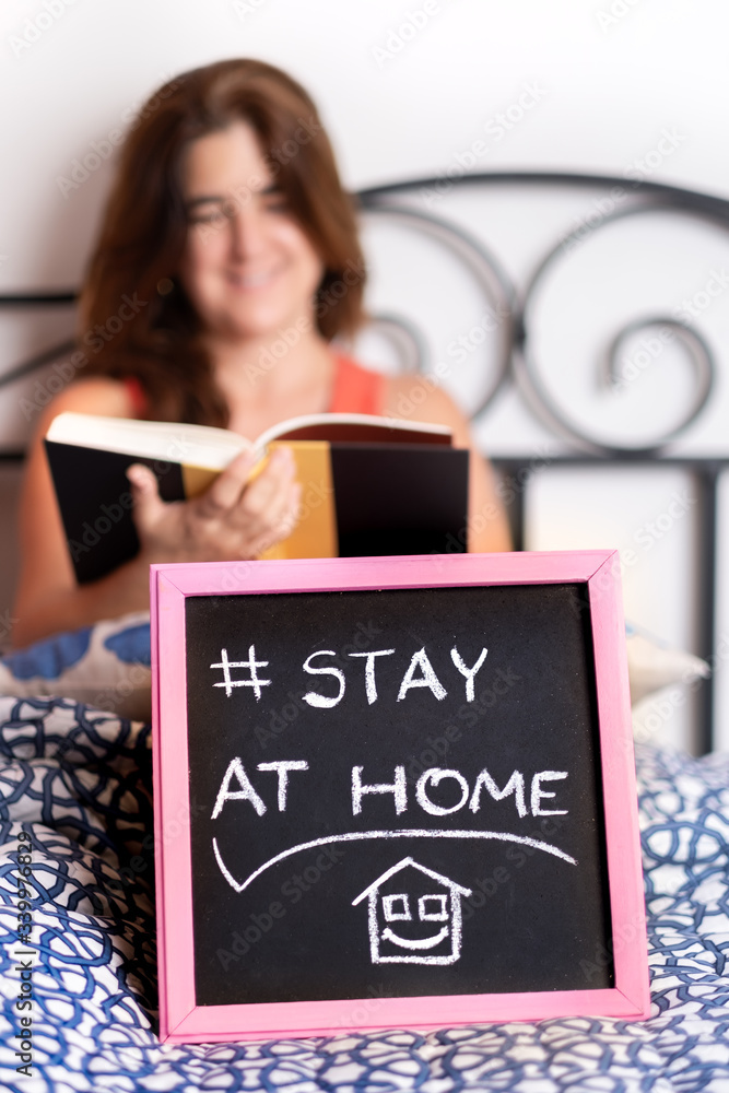 Woman in her bedroom reading during a quarantine with a Stay At Home sign - Focused on the sign Woman in her bedroom reading during a quarantine with a Stay At Home sign - Focused on the sign