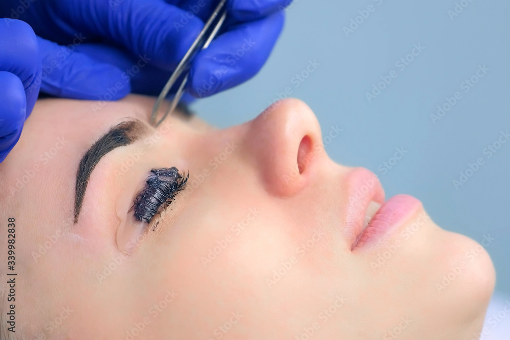 Beautician plucking eyebrows with tweezers to woman in beauty salon during tint eyebrow and lash laminating procedure, side view. Cosmetologist shaping girl's eyebrows in cosmetology clinic.
