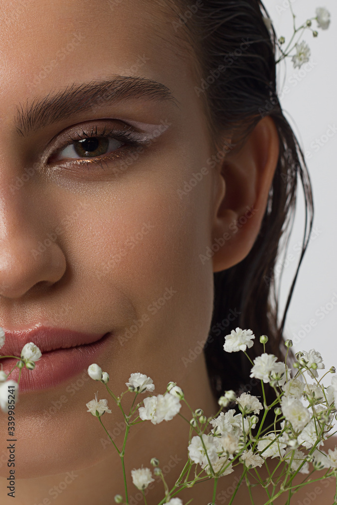 Fototapeta premium Close up half portrait of a face with beautiful skin and nude makeup, eyeliner and long eyelashes, puffy natural lips and white flowers. Fashion photography. Cosmetology and spa. Freshness of spring