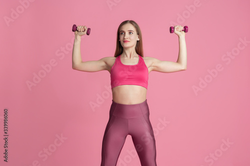 Confidence. Beautiful young female athlete practicing in studio, monochrome pink portrait. Sportive fit caucasian model with weights. Body building, healthy lifestyle, beauty and action concept.