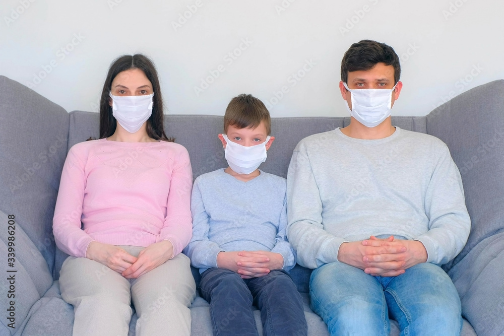 Mom, dad and son in protective medical masks sitting on couch at home and looking at camera, coronavirus pandemic quarantine. Worldwide global COVID-19 epidemic. Fighting with 2019-ncov infection.