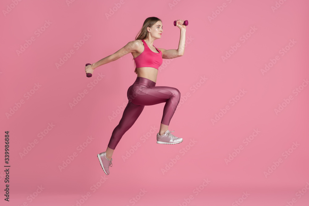 In jump, flight. Beautiful young female athlete practicing in studio, monochrome pink portrait. Sportive fit caucasian model with weights. Body building, healthy lifestyle, beauty and action concept.