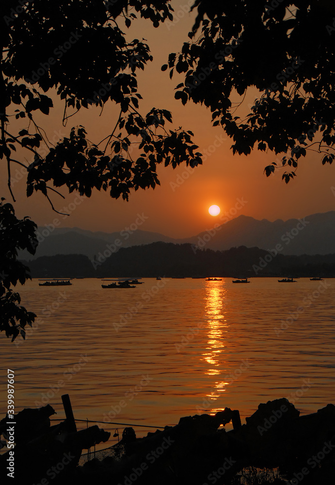 West Lake (Xi Hu) sunset in Hangzhou, Zhejiang Province, China. View of West Lake in Hangzhou at sunset with an orange light across the lake with boats and hills. Framed by a tree and lilies.