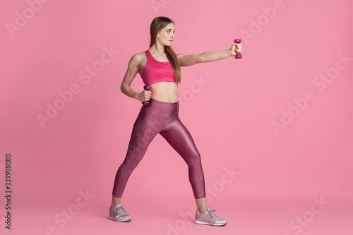 Active. Beautiful young female athlete practicing in studio, monochrome pink portrait. Sportive fit caucasian model with weights. Body building, healthy lifestyle, beauty and action concept.