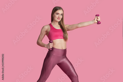 Active. Beautiful young female athlete practicing in studio, monochrome pink portrait. Sportive fit caucasian model with weights. Body building, healthy lifestyle, beauty and action concept.