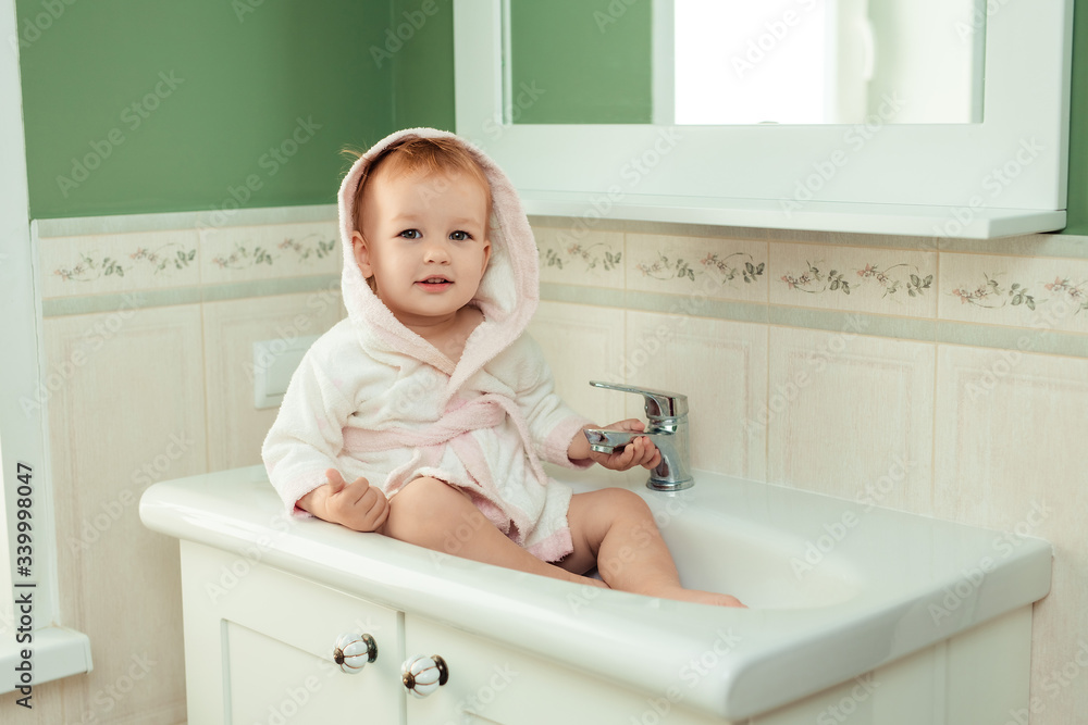 baby bathing and hygiene. care for young children. cute charming girl 1 year old in a white bathrobe sits in the sink in the bathroom baby bathing and hygiene. care for young children. cute charming girl 1 year old in a white bathrobe sits in the sink in the bathroom