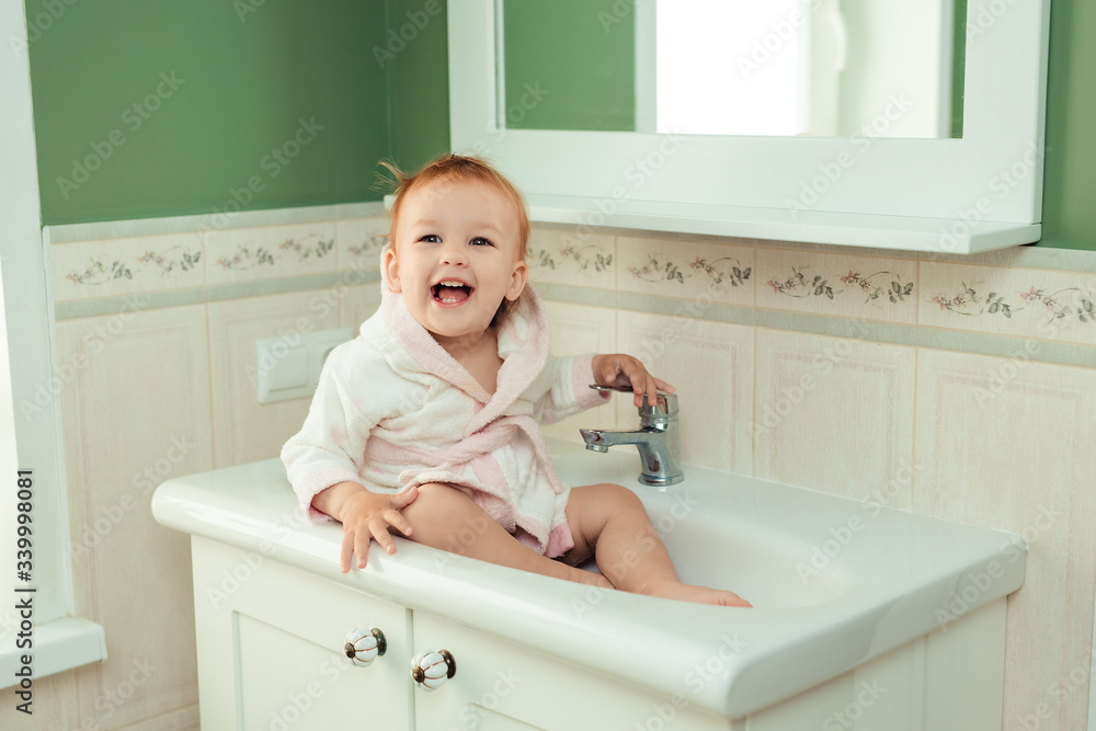 baby bathing and hygiene. care for young children. cute charming girl 1 year old in a white bathrobe sits in the sink in the bathroom baby bathing and hygiene. care for young children. cute charming girl 1 year old in a white bathrobe sits in the sink in the bathroom