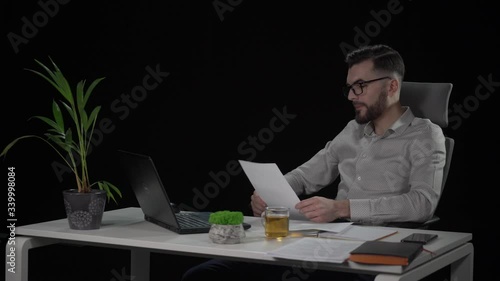 Copywriter reviews article from papers, crumbles and throws out bad sheet. Bearded young man in eyeglasses at white desk. Indoor studio shot on black background.