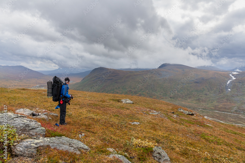 Man backpack hiker at Kungsleden trail admiring nature of Sarek in Sweden Lapland with mountains, rivers and lakes, birch and spruce tree forests. Early autumn colors in stormy weather