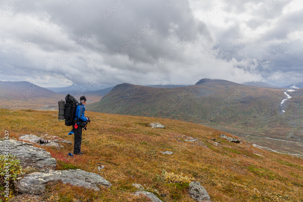 Man backpack hiker at Kungsleden trail admiring nature of Sarek in Sweden Lapland with mountains, rivers and lakes, birch and spruce tree forests. Early autumn colors in stormy weather Man backpack hiker at Kungsleden trail admiring nature of Sarek in Sweden Lapland with mountains, rivers and lakes, birch and spruce tree forests. Early autumn colors in stormy weather