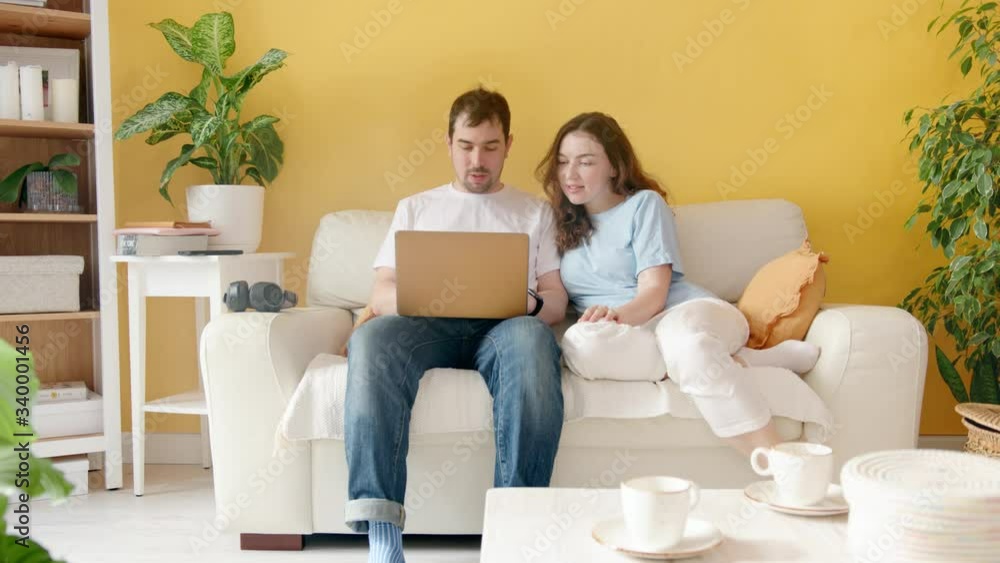 A Couple Makes Online Shopping while Sitting on the Couch. A Man and a Woman Work Together on a Laptop. Cozy Home Interior. A Couple Makes Online Shopping while Sitting on the Couch. A Man and a Woman Work Together on a Laptop. Cozy Home Interior.