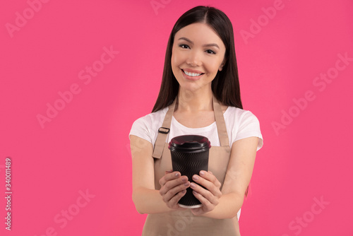 Nice stylish brunette asian girl, barista in casual tshirt and apron, offers a cup of coffee or tea, isolated over pink background