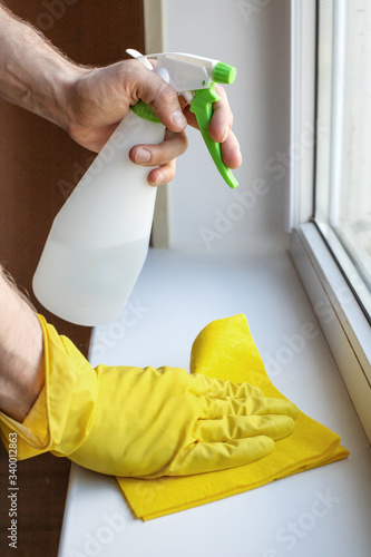 the hand of a man in a yellow rubber glove wipes the white window sill and window glass with a yellow viscose cloth using an antiseptic spray in a white color