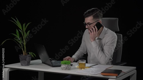 Bearded young businessman during working on laptop gets phone call, turns off laptop and leaves workplace. Indoor studio shot on black background.