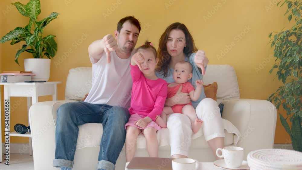 A Young Family, Father, Mother and Two Children Together Give a Thumbs Down. Cozy Interior with Yellow Wall, White Couch and House Plants. A Young Family, Father, Mother and Two Children Together Give a Thumbs Down. Cozy Interior with Yellow Wall, White Couch and House Plants.